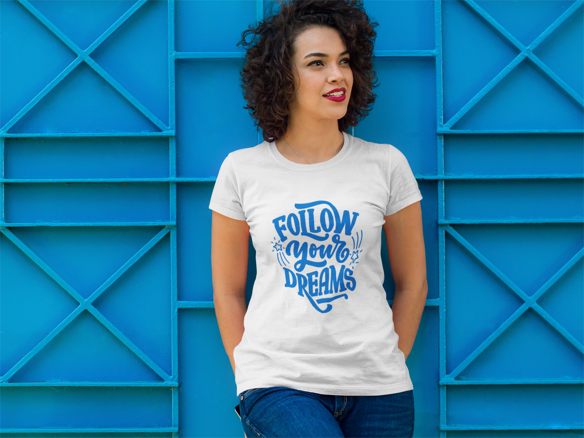 Curly hair woman standing in front of blue wall wearing a follow your dreams t-shirt.