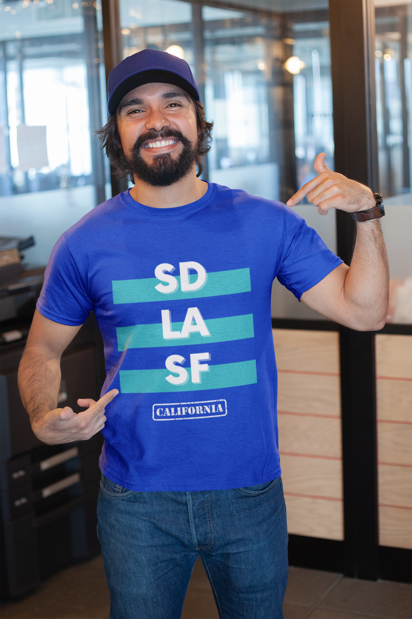 Bearded guy pointing at his blue SD LA SF t-shirt inside of office buiding.