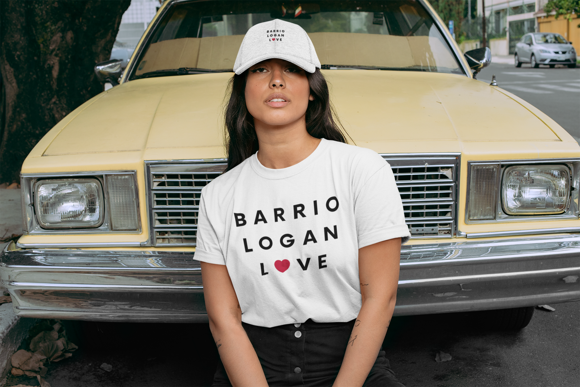 Pretty Latina posing in front of a lowrider wearing matching Barrio Logan t-shirt and hat.