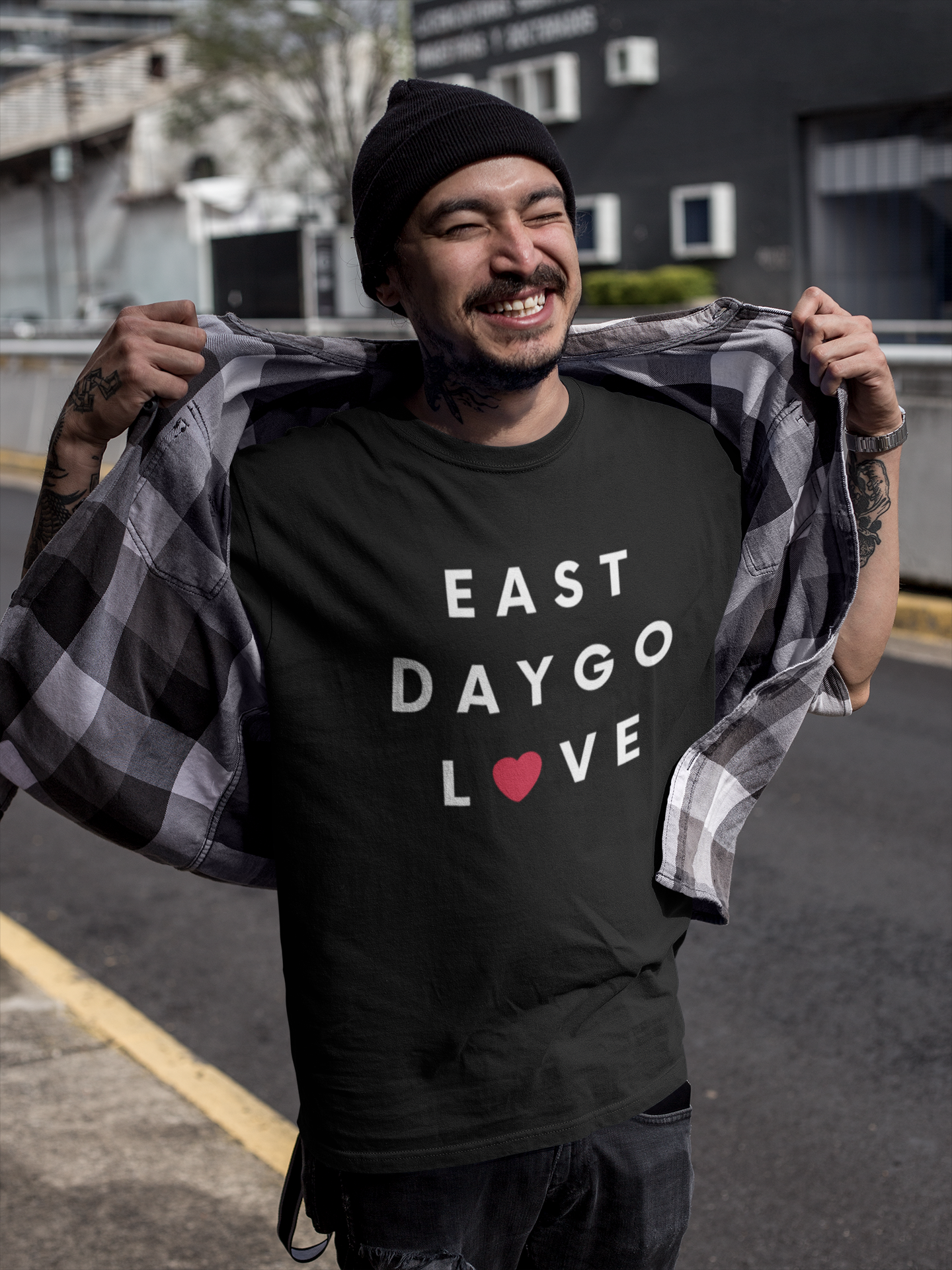 Smiling Asian man with tattoos standing outside buildings wearing a beanie and East Daygo t-shirt.