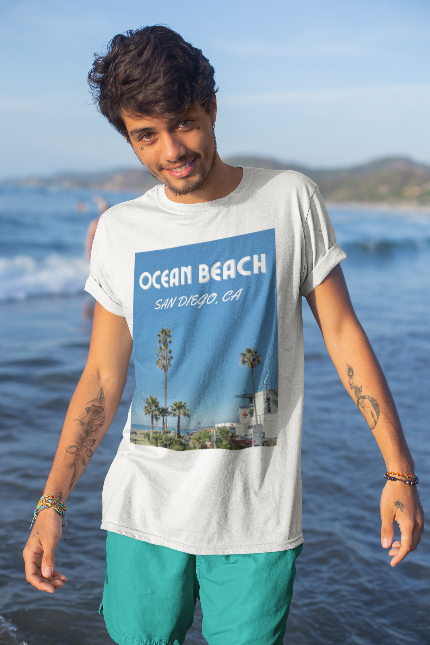 Smiling guy with tattoos wearing Ocean Beach t-shirt and green shorts posing in front on waves.