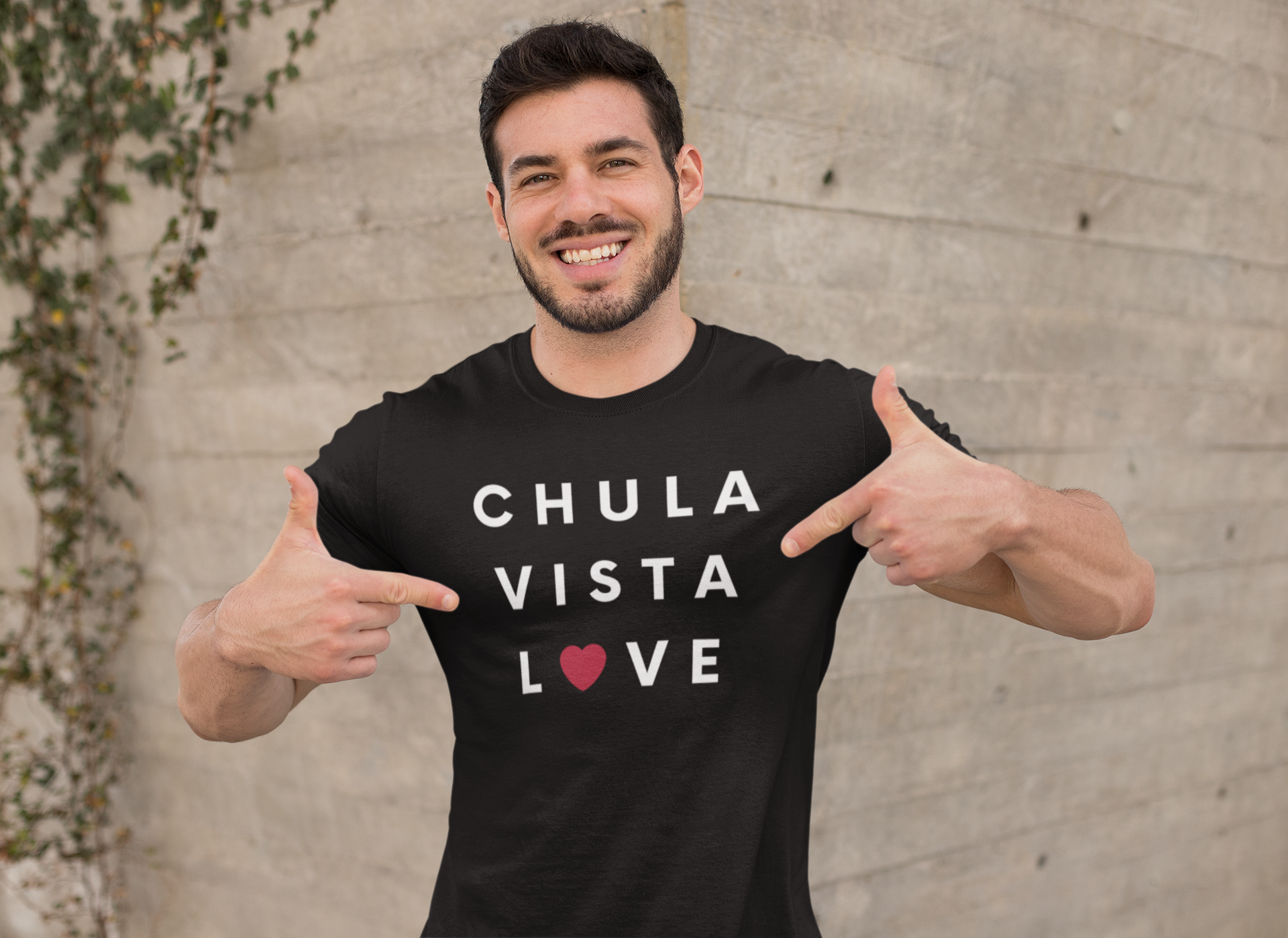 Happy Hispanic man standing in front of brick wall pointing at his Chula Vista t-shirt.