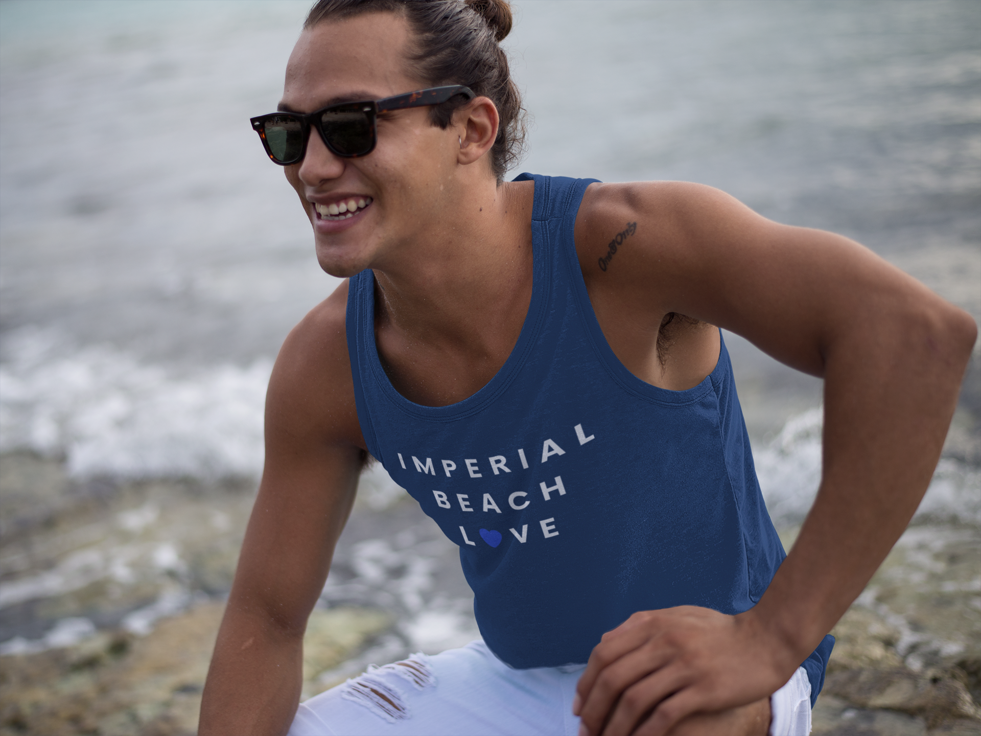 Young man with man-bun wearing sunglasses and Imperial Beach tank top kneeling down in front of ocean waves.