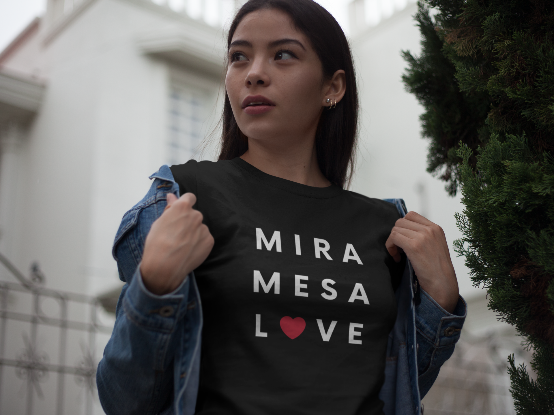 Pretty Asian woman standing outside a home with her jacket half-way off showing her Mira Mesa t-shirt.