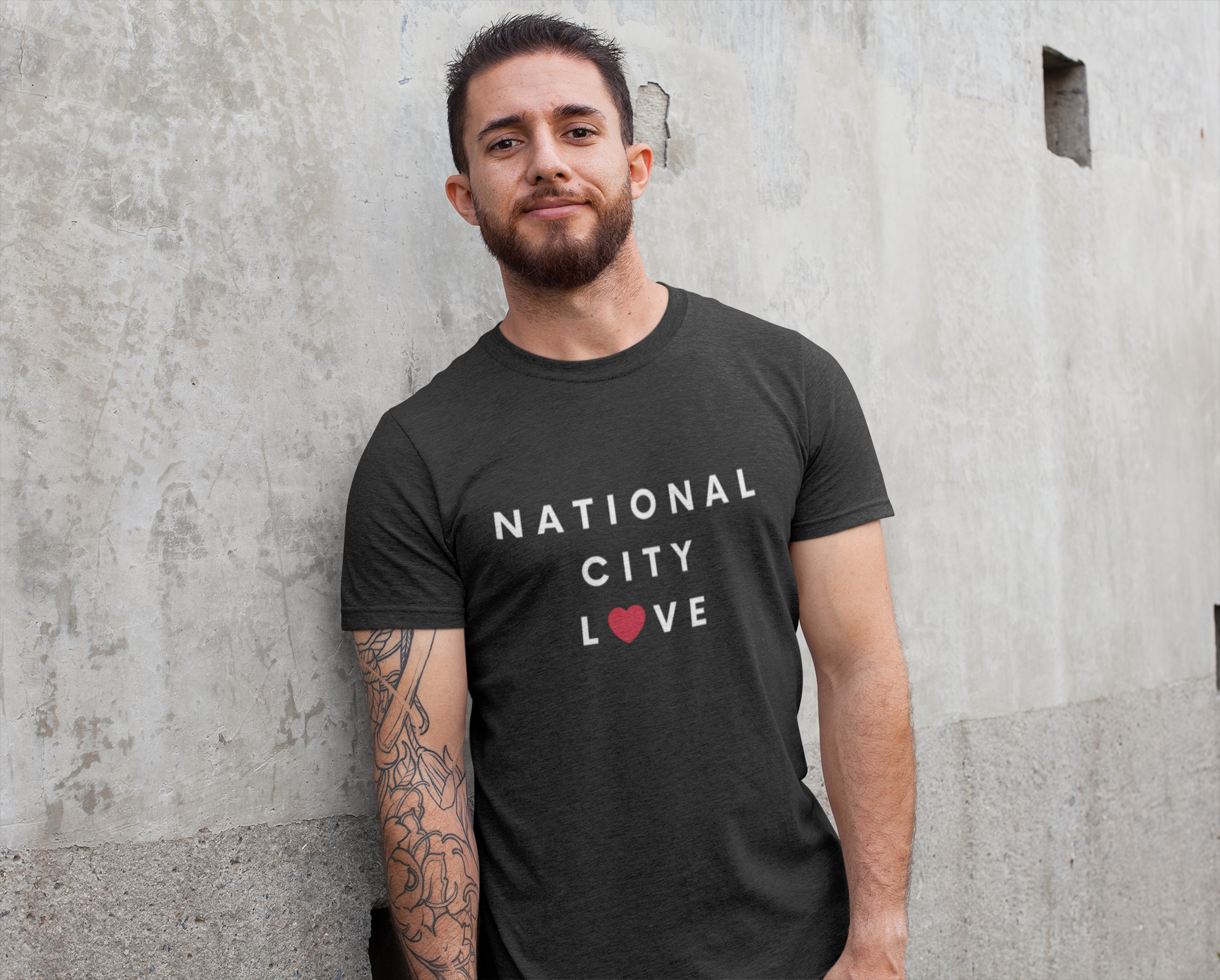 Tattooed guy standing again concrete wall wearing a National City t-shirt.